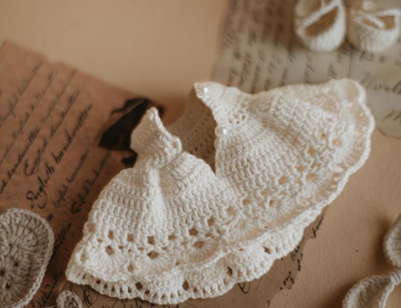 May include: A white crocheted baby dress with a ruffled hem and a pearl button detail.