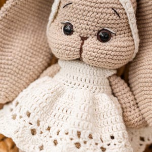 May include: A crocheted bunny toy wearing a white dress with a lacy trim. The bunny has brown eyes and a beige body.