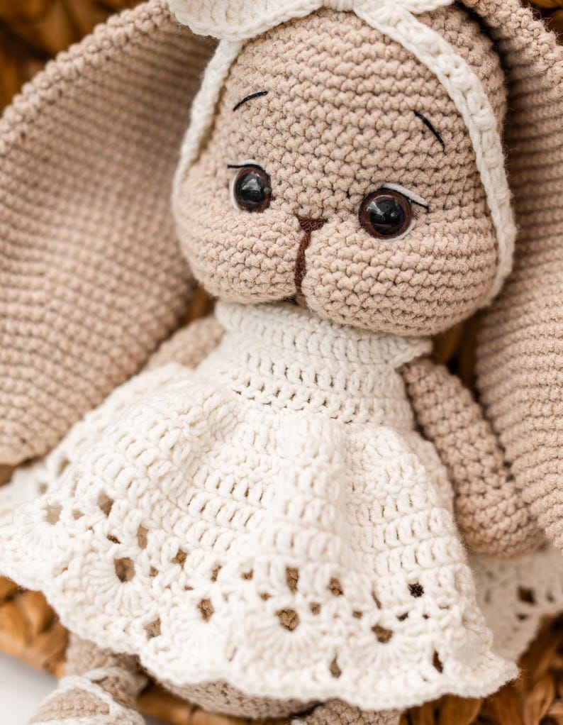 May include: A crocheted bunny toy wearing a white dress with a lacy trim. The bunny has brown eyes and a beige body.