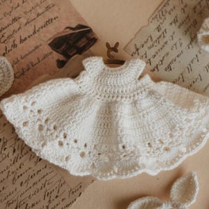 May include: A white crocheted baby dress with a delicate lace pattern. The dress has a simple, sleeveless design and a flared skirt.