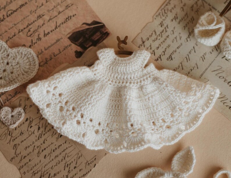 May include: A white crocheted baby dress with a delicate lace pattern. The dress has a simple, sleeveless design and a flared skirt.