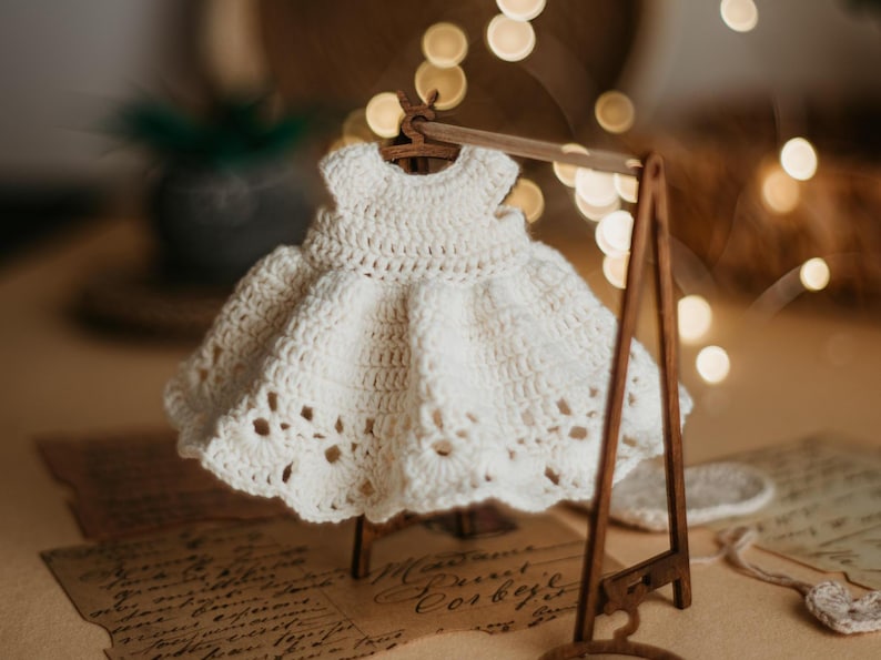 May include: A white crocheted dress hangs on a wooden clothes rack. The dress has a delicate lace trim and is displayed against a backdrop of blurred lights.