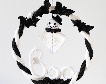 Könnte beinhalten: Ein handgefertigter Halloween-Kranz mit einem schwarz-weißen, gedrehten Seil-Design. Der Kranz ist mit schwarzen Fledermaus-Ausschnitten und einem gehäkelten Geist mit Hut verziert. Das Wort "Boo" ist aus weißem Garn geschrieben und sorgt für eine festliche Note.