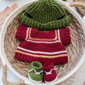 Puede incluir: Un conjunto de bebé hecho a mano con ganchillo en una cesta tejida. El conjunto incluye un gorro verde, un suéter rojo con rayas blancas y amarillas, y botines a juego verdes y rojos con cordones blancos. Los artículos están hechos de hilo suave.