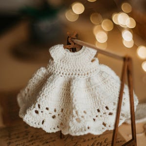 May include: A white crocheted dress hangs on a wooden clothes rack. The dress has a delicate lace pattern and a full skirt. The dress is displayed on a vintage-looking background.