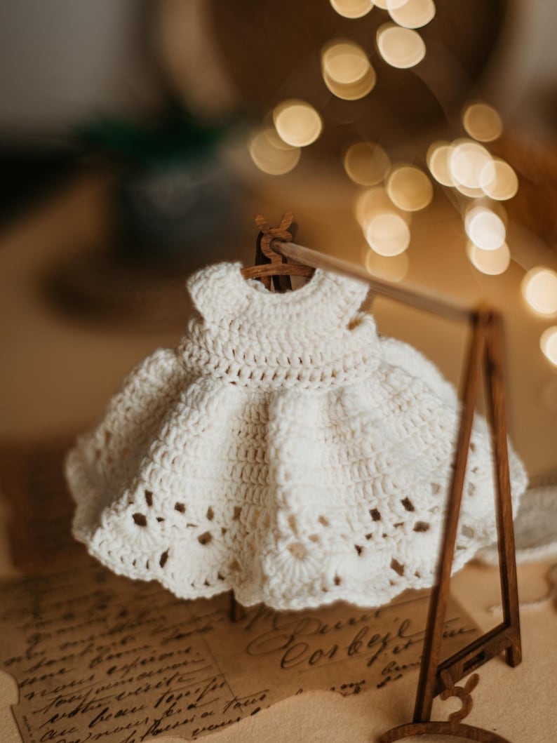 May include: A white crocheted dress hangs on a wooden clothes rack. The dress has a delicate lace pattern and a full skirt. The dress is displayed on a vintage-looking background.