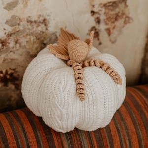 Op de afbeelding: Een witte gehaakte pompoen met een bruine gehaakte steel en bladeren. De pompoen ligt op een gestreepte stof.