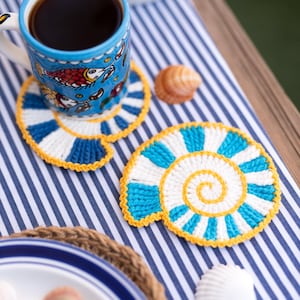 Pode incluir: Uma caneca de cerâmica azul com um desenho de peixe está sobre um descanso de copo listrado azul e branco. O descanso de copo é feito de crochê em um padrão espiral com listras azuis e brancas e uma borda amarela. Uma concha e um prato também são visíveis.