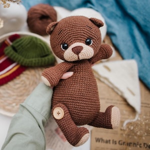 Può includere: Un orsacchiotto di peluche marrone all'uncinetto con occhi neri e un bottone marrone sulla sua zampa. L'orso è tenuto da una mano.