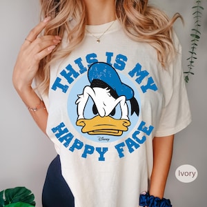 May include: Ivory-colored t-shirt featuring a distressed graphic of Donald Duck with the text "THIS IS MY HAPPY FACE" in blue. The Disney logo is visible below the character. The shirt has short sleeves.