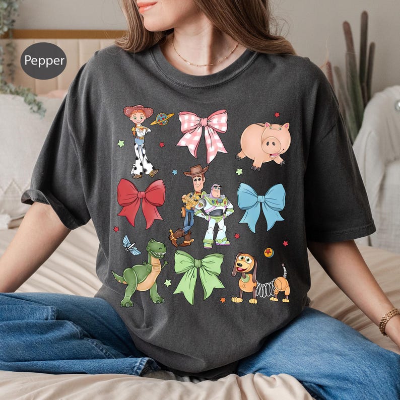Op de afbeelding: Donkergrijs T-shirt met stripfiguren en strikken. Het shirt heeft illustraties van een varken, een dinosaurus, een hond en andere karakters. Het woord "Pepper" staat in een cirkel linksboven op het shirt.