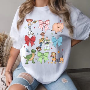 Op de afbeelding: Wit T-shirt met kleurrijke illustraties van personages en strikken. Het ontwerp bevat een roze varken, een groene dinosaurus en een teckel, samen met andere personages en strikken. Het shirt is een casual kledingstuk.