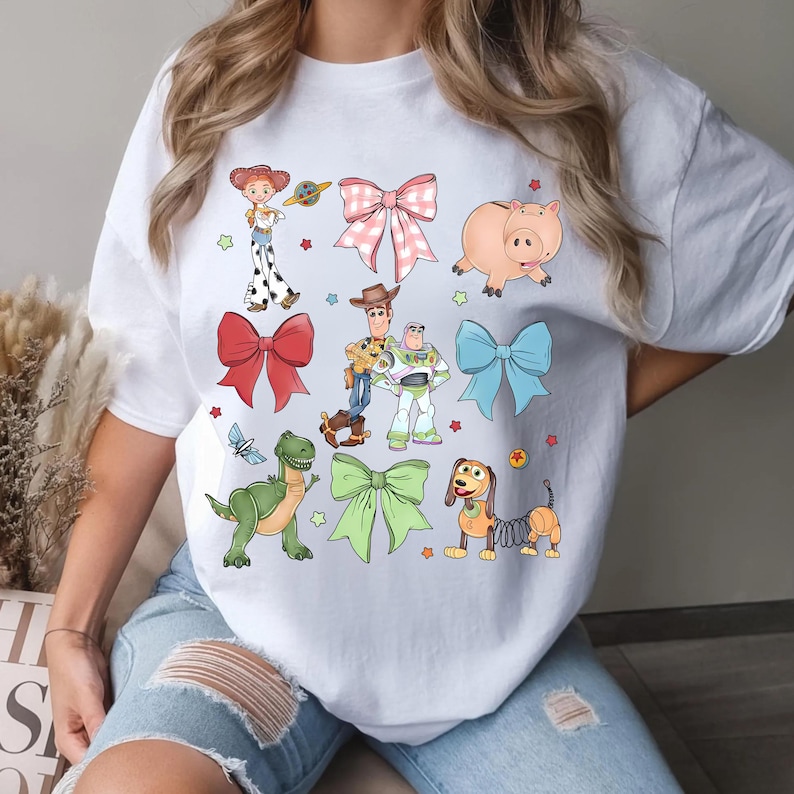 Op de afbeelding: Wit T-shirt met kleurrijke illustraties van personages en strikken. Het ontwerp bevat een roze varken, een groene dinosaurus en een teckel, samen met andere personages en strikken. Het shirt is een casual kledingstuk.