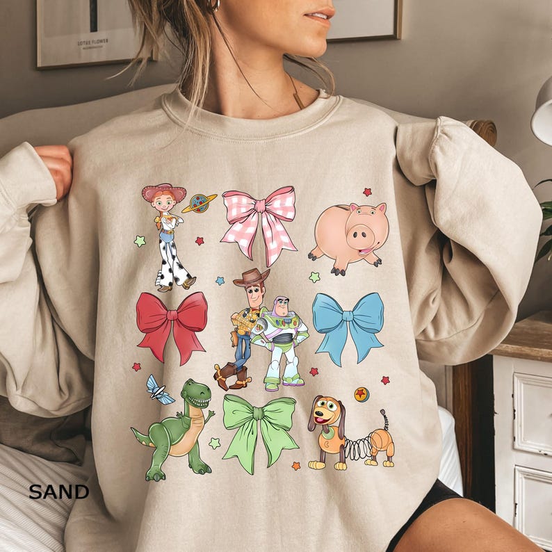 Op de afbeelding: Beige sweatshirt met kleurrijke cartoonfiguren en strikken. Het ontwerp bevat Woody, Buzz Lightyear en Jessie, een roze varken, een groene dinosaurus en een teckel. Het woord "SAND" staat onderaan afgedrukt.