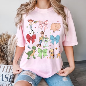 May include: A pale pink t-shirt with cartoon characters and bows. The design includes images of a pig, a dinosaur, a dog, and other characters. The shirt also has several bows in different colours and patterns. The shirt is made of a soft material.