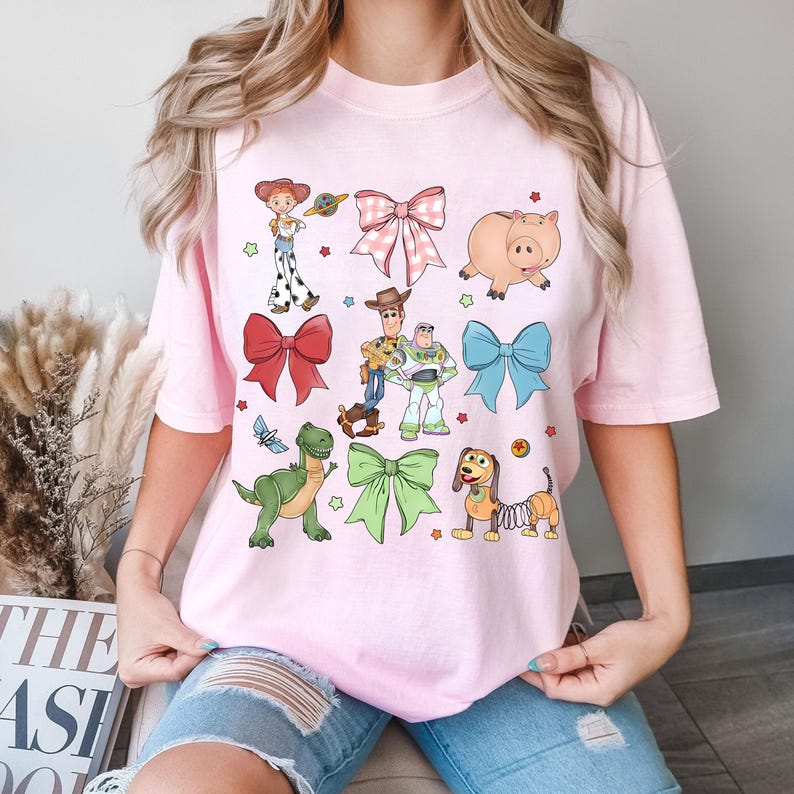 Op de afbeelding: Een lichtroze T-shirt met stripfiguren en strikken. Het ontwerp bevat afbeeldingen van een varken, een dinosaurus, een hond en andere personages. Het shirt heeft ook verschillende strikken in verschillende kleuren en patronen. Het shirt is gemaakt van zacht materiaal.