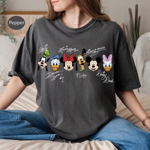May include: Dark grey t-shirt featuring cartoon character faces of Goofy, Donald Duck, Minnie Mouse, Pluto, Mickey Mouse, and Daisy Duck. Each character's name is written in a cursive font above their face. The shirt has a relaxed fit.
