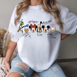 May include: White t-shirt with cartoon images of Disney characters, including Goofy, Donald Duck, Minnie Mouse, Pluto, Mickey Mouse, and Daisy Duck. Each character has a signature above or below their image. The shirt is a casual style.