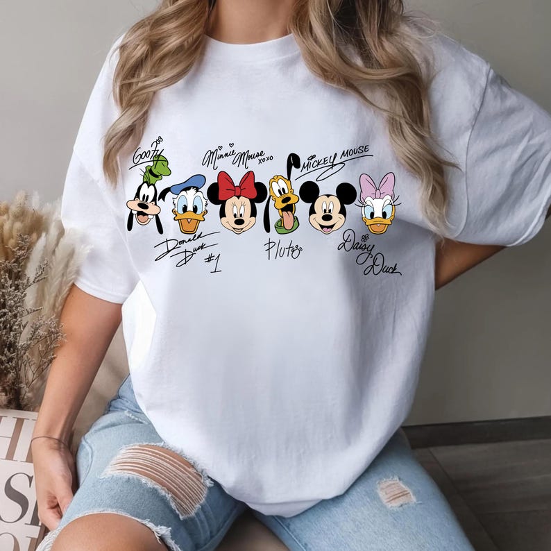 May include: White t-shirt with cartoon images of Disney characters, including Goofy, Donald Duck, Minnie Mouse, Pluto, Mickey Mouse, and Daisy Duck. Each character has a signature above or below their image. The shirt is a casual style.