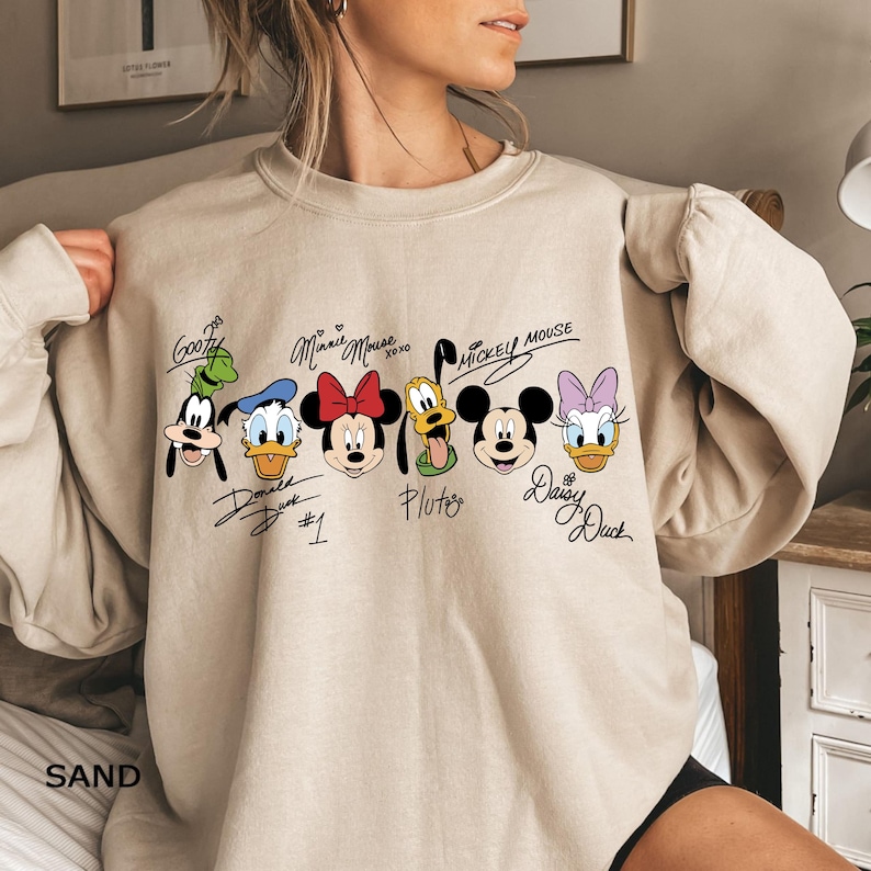 May include: A sand-coloured sweatshirt with a colourful graphic of Disney characters, including Goofy, Donald Duck, Minnie Mouse, Pluto, Mickey Mouse, and Daisy Duck. Each character has their name written above them.