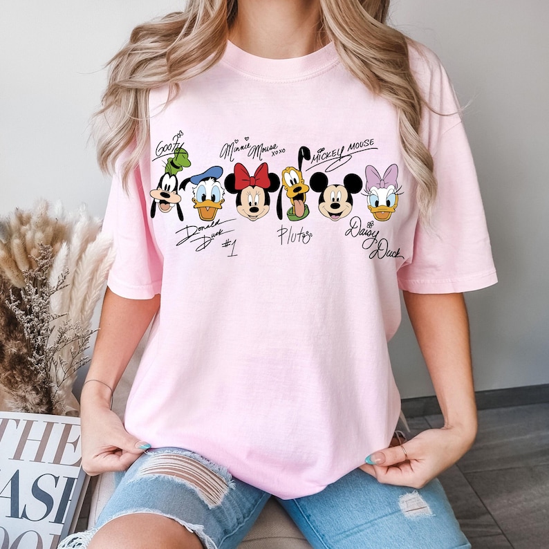 May include: A pale pink t-shirt displaying cartoon character faces and signatures. Characters include Goofy, Donald Duck, Minnie Mouse, Pluto, Mickey Mouse, and Daisy Duck. The shirt is a relaxed, casual style.