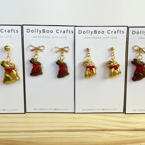May include: Four pairs of handmade bunny rabbit earrings. Two pairs are gold with red bows, and two pairs are brown with red bows. The earrings are displayed on white cards with the text "DollyBoo Crafts Handmade with Love" and a website address.