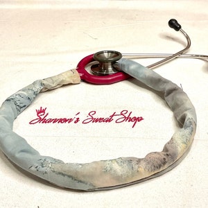 May include: A stethoscope with a gray and white patterned fabric cover. The stethoscope is wrapped around the fabric cover, forming a circle. The text "Shannon's Sweet Shop" is embroidered in pink on the fabric.