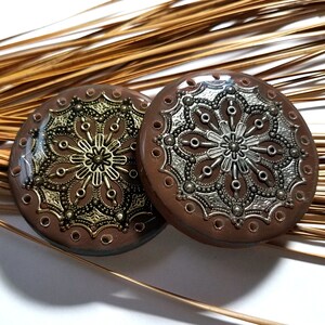 May include: Two round, brown, decorative coasters with intricate gold and silver filigree designs. The coasters have a raised edge and a circular pattern of holes around the edge.