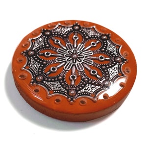 May include: A round, orange, decorative object with a silver, ornate, mandala design in the center. The object has small holes around the edge.