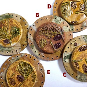 May include: A collection of five ceramic pieces featuring a nature theme. Each piece has a circular design with a central raised image of pine needles and pine cones in shades of green, brown, and tan. The outer ring is a lighter brown with small holes.