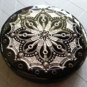 May include: A round, black button with a silver floral design. The button has a glossy finish and a detailed, symmetrical pattern. Small holes are visible around the edge of the button, adding to its decorative appeal. This button is suitable for sewing projects.