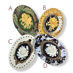 May include: Four oval-shaped cameo brooches. Each brooch features a central cameo design with floral motifs. The brooches have different backgrounds, including green, yellow, white, and black, with ornate frames and decorative details.