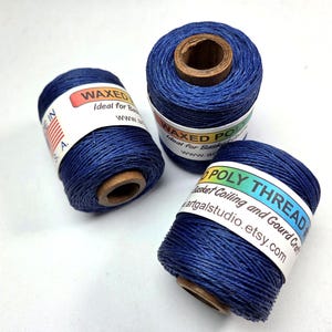 May include: Three spools of dark blue waxed poly thread. Each spool has a wooden core and a label with text. The labels read "WAXED POLY THREAD" and "Basket Coiling and Gourd Craft".