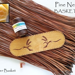 May include: A Pine Needle BASKET KIT featuring a bundle of long brown pine needles, a spool of brown waxed poly thread, a light wood oval base with two wheat stalk engravings, and a small hexagonal piece of beeswax for basket crafting.