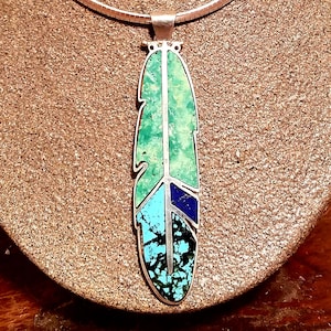 Könnte beinhalten: Eine silberne Halskette mit einem Anhänger in Form einer Feder aus Türkis und Lapislazuli. Die Feder besteht aus verschiedenen Farbtönen von Türkis und Lapislazuli und ist in Silber gefasst.