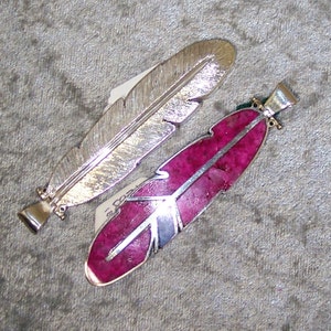 May include: Two silver feather pendants, one with a smooth silver finish and the other with a pink stone inlay.