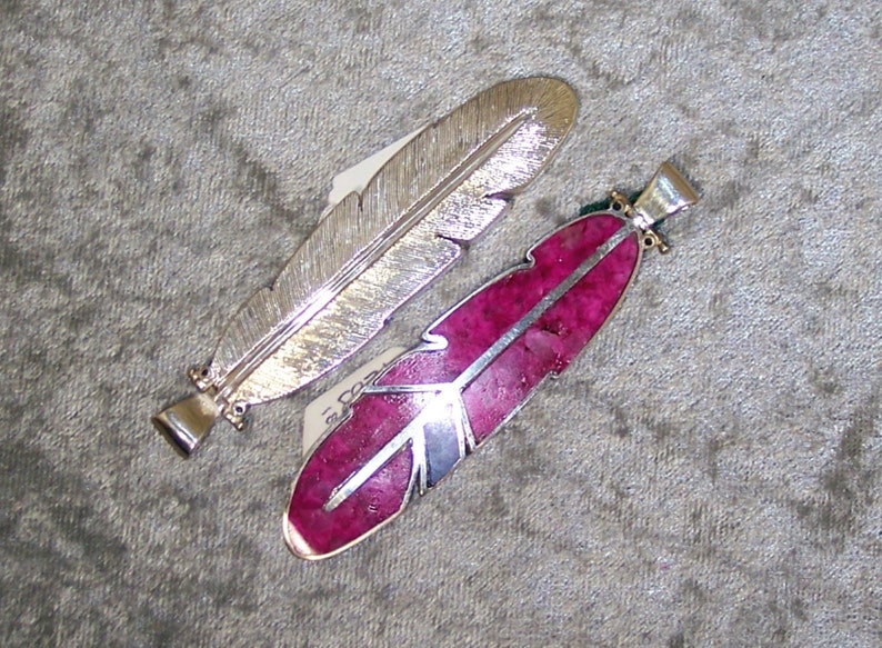 May include: Two silver feather pendants, one with a smooth silver finish and the other with a pink stone inlay.
