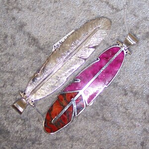 K&ouml;nnte beinhalten: Zwei silberne Federn Anh&auml;nger, einer mit einem rosa und orangefarbenen Stein-Inlay und der andere mit einer silbernen Oberfl&auml;che.