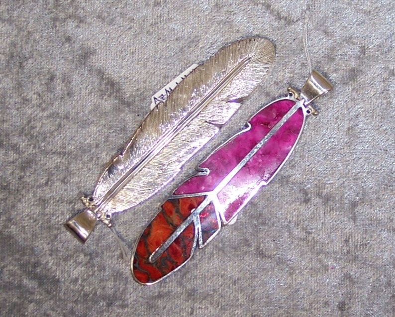 K&ouml;nnte beinhalten: Zwei silberne Federn Anh&auml;nger, einer mit einem rosa und orangefarbenen Stein-Inlay und der andere mit einer silbernen Oberfl&auml;che.