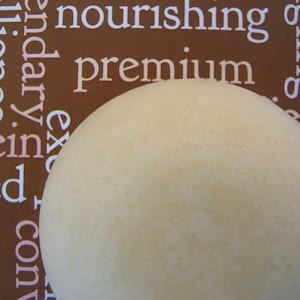 May include: A close-up of a round, off-white solid bar of soap. The soap is sitting on a brown surface with white text.