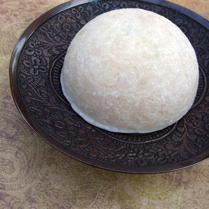 May include: A white, round, cake-like dessert sits on a decorative, dark brown, metal plate with intricate floral designs.