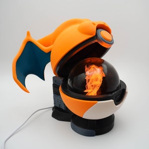 May include: An orange and white lamp shaped like a Pokeball with a glowing orange figure inside. The lamp has a blue wing on the side.