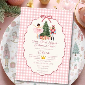 May include: A rectangular invitation card with a pink and white gingham border. The center features a watercolor illustration of a ballerina, a Christmas tree, and a nutcracker. Text reads: Our Little Sugar Plum is One! Clara, December 10.