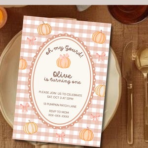 May include: A rectangular invitation card featuring a light pink and white gingham background with illustrations of small orange pumpkins and pink bows. The center has an oval frame with the text: Oh my Gourd! Olive is turning one. PLEASE JOIN US TO CELEBRATE SAT, OCT 3 AT 12PM 13 PUMPKIN PATCH LANE RSVP TO MOM XXX-XXX-XXXX.