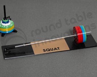May include: A miniature weightlifting set with a barbell and weight plates in red, blue, and green. The barbell rests on a black platform with the word "SQUAT" printed on it. A smaller weight set is also present.