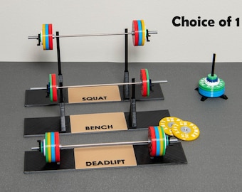 May include: Three miniature weightlifting stands with black bases and silver bars. The stands are labelled "Squat", "Bench", and "Deadlift". Each stand has a set of colourful weight plates. A separate stand holds a stack of yellow weight plates with the text "Power Lift" printed on them.