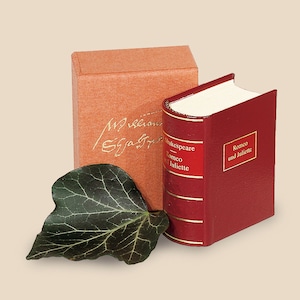 May include: A small red leather-bound book with gold accents and the title "Romeo and Juliet" by Shakespeare. The book is sitting on a brown box with a handwritten inscription.