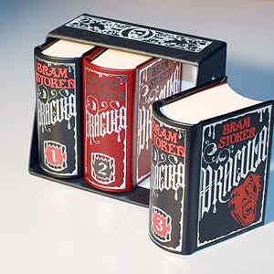 May include: A set of three black and red hardback books with the title "Dracula" by Bram Stoker. The books are numbered 1, 2, and 3.