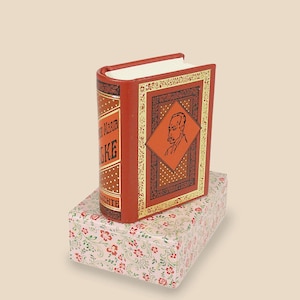 May include: A small red book with gold accents and a portrait of a man on the cover. The book is sitting on a pink box with a floral pattern.