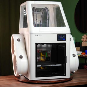 Könnte beinhalten: Ein weißer Snapmaker 3D-Drucker mit einem transparenten Gehäuse, einem digitalen Touchscreen-Display an der Vorderseite und zwei seitlich montierten runden Modulen. Das Gerät hat ein modulares Design mit sichtbaren internen Komponenten.